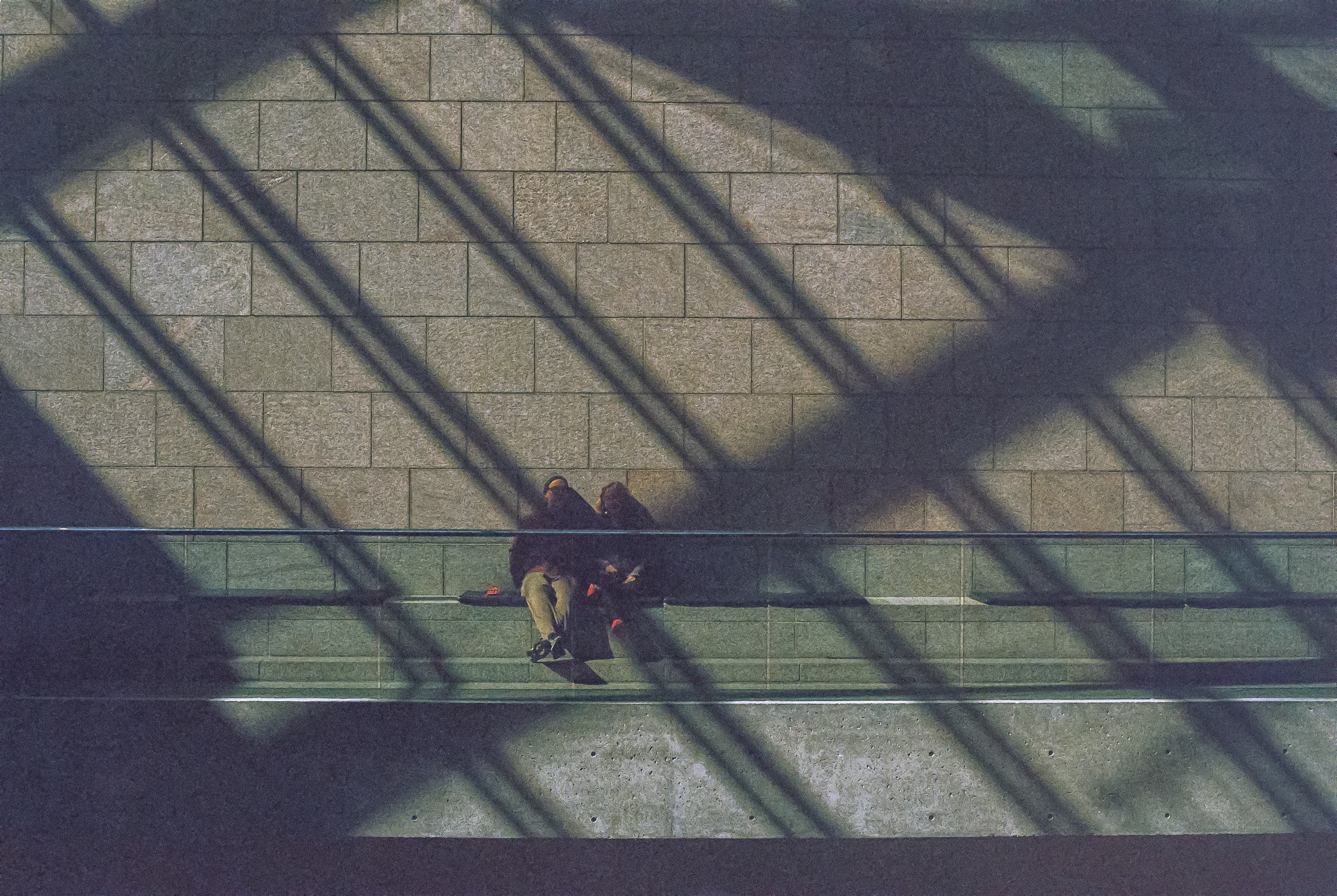 Two people sitting on a concrete bench, with shadows from the glass roof and its structure arcing across them diagonally.